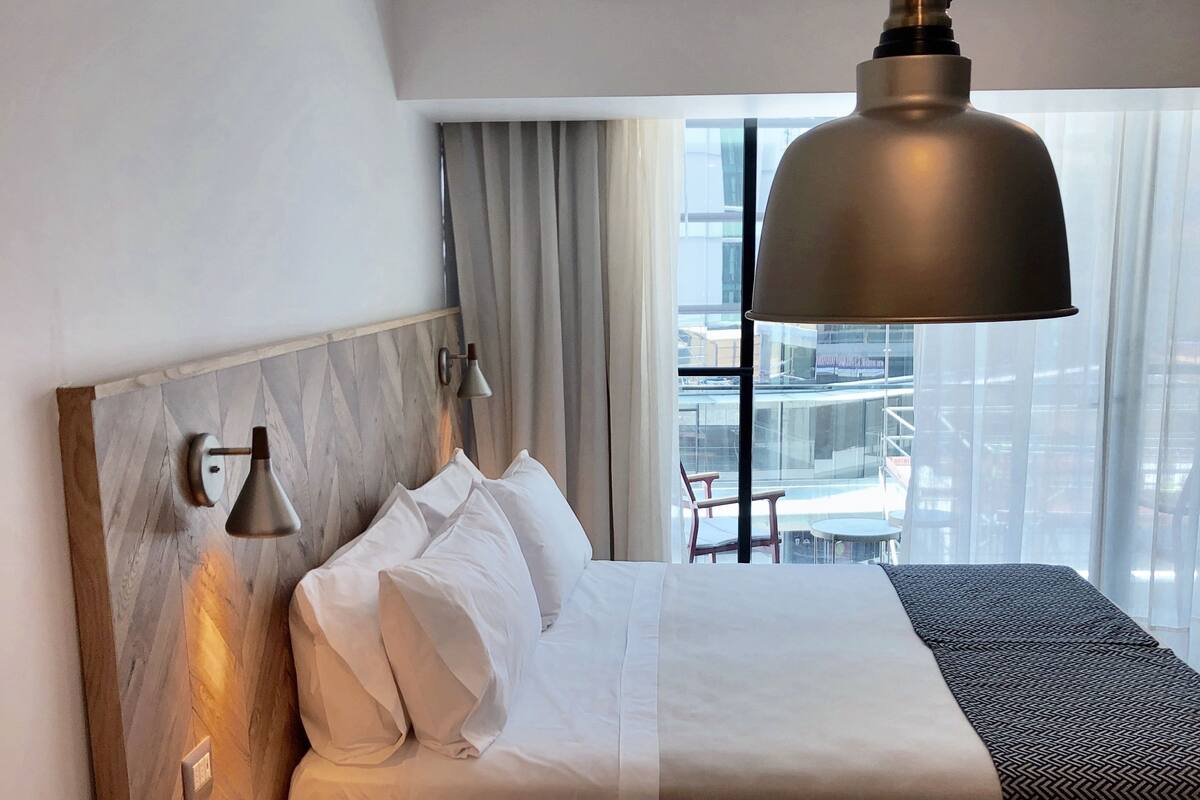 Habitación de hotel con una cama, una mesita de noche con productos de desayuno, una lámpara y vistas a la ciudad a través de amplias ventanas.