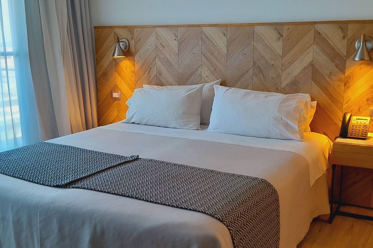 Habitación de hotel con una cama grande, dos lámparas de noche, una mesita de noche con un teléfono y un cabecero con estampado de espiga.