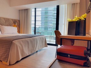 A hotel room with a bed, a desk with a chair, a suitcase, and a view of buildings through large windows.