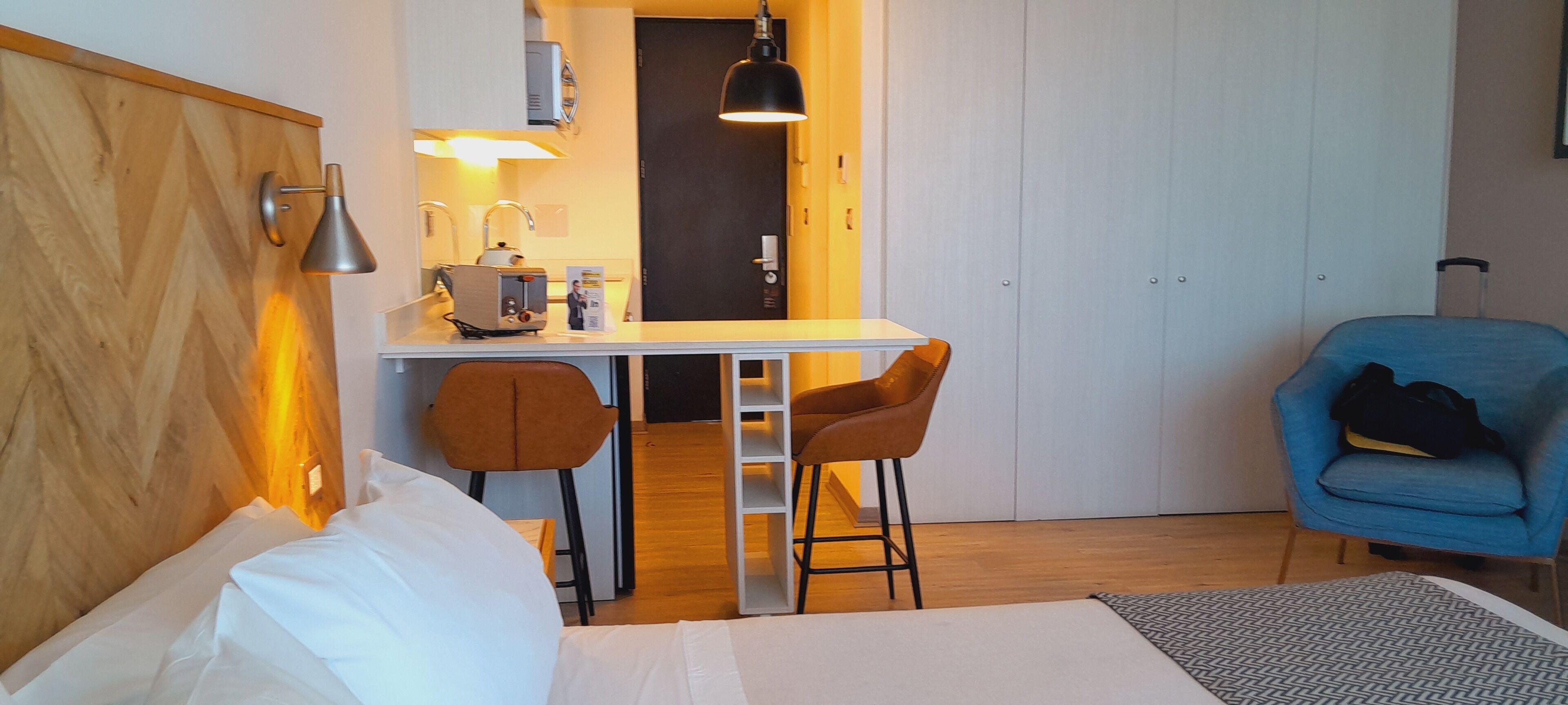 A hotel room with a bed, a desk with two chairs, a blue armchair, and a wardrobe.
