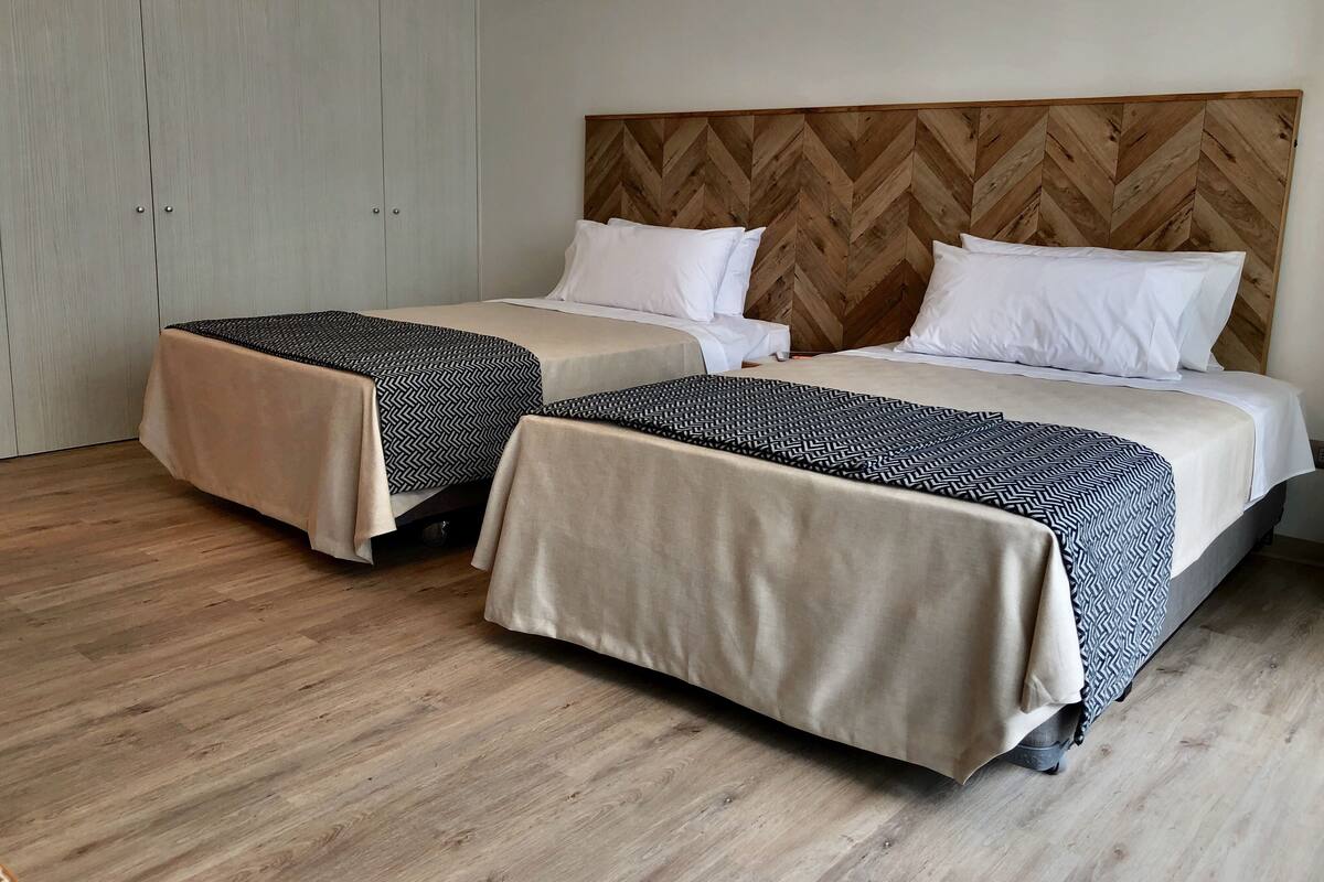 Habitación de hotel con dos camas, cada una con colcha y almohadas estampadas, cabecero de madera con patrón de espiga y armario con acabado claro.