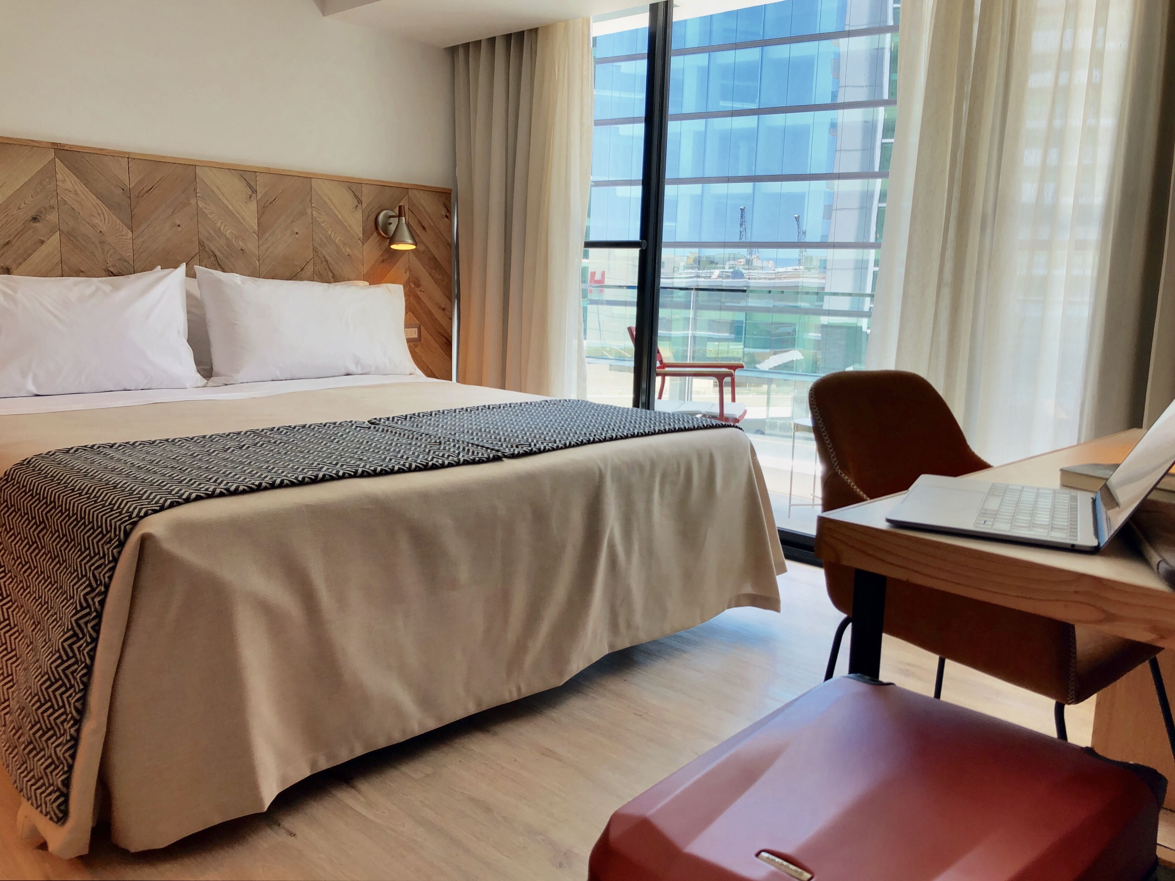 Habitación de hotel con una cama grande, un escritorio con una computadora portátil, una silla y vistas a un edificio moderno a través de amplios ventanales.