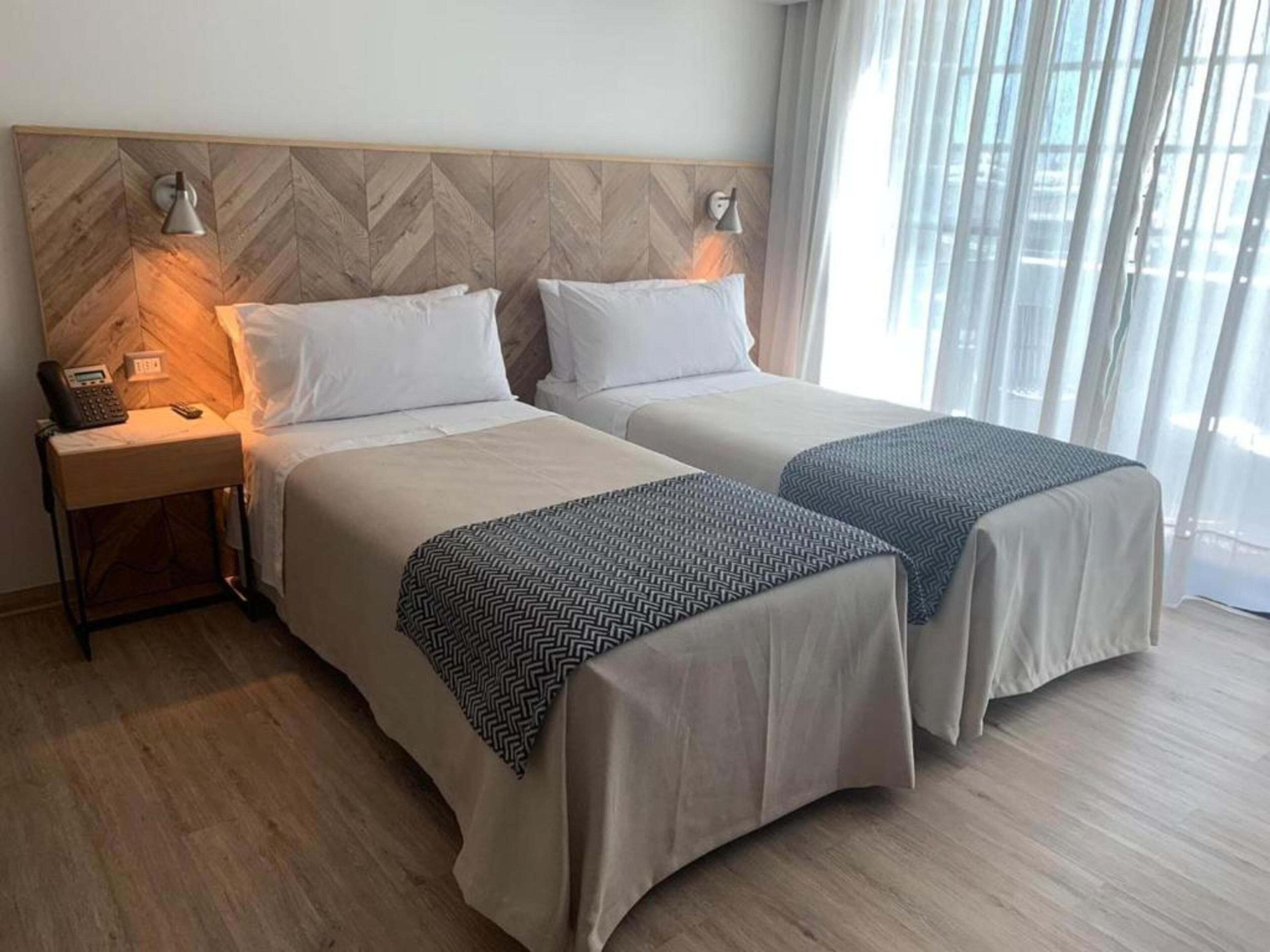 Een hotelkamer met twee bedden, een houten hoofdeinde, bedlampen, een telefoon en een raam met gordijnen.