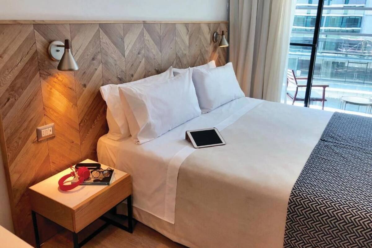 Habitación de hotel con cama, mesita de noche, lámpara y vista a un edificio.