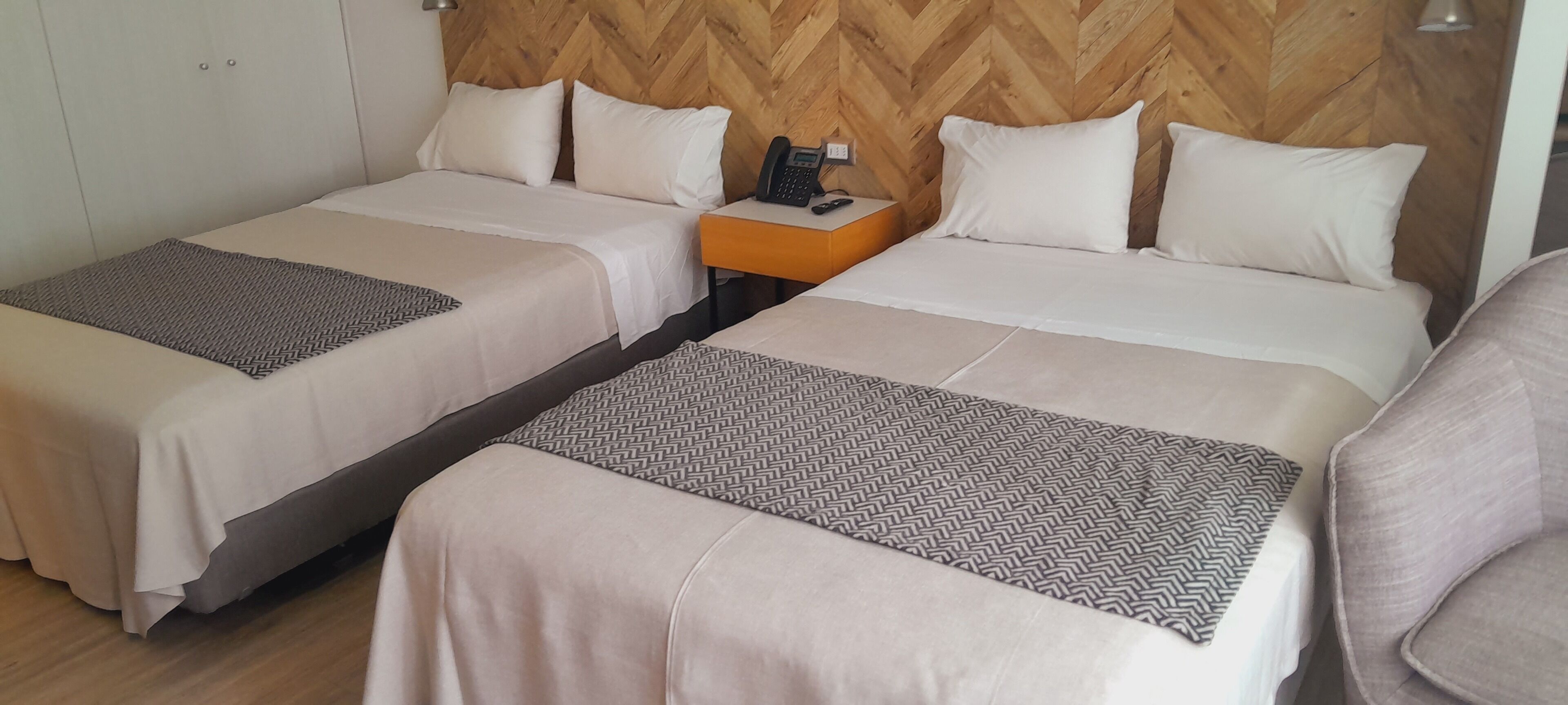 Habitación de hotel con dos camas, cabecera de madera, mesita de noche con teléfono y una silla.