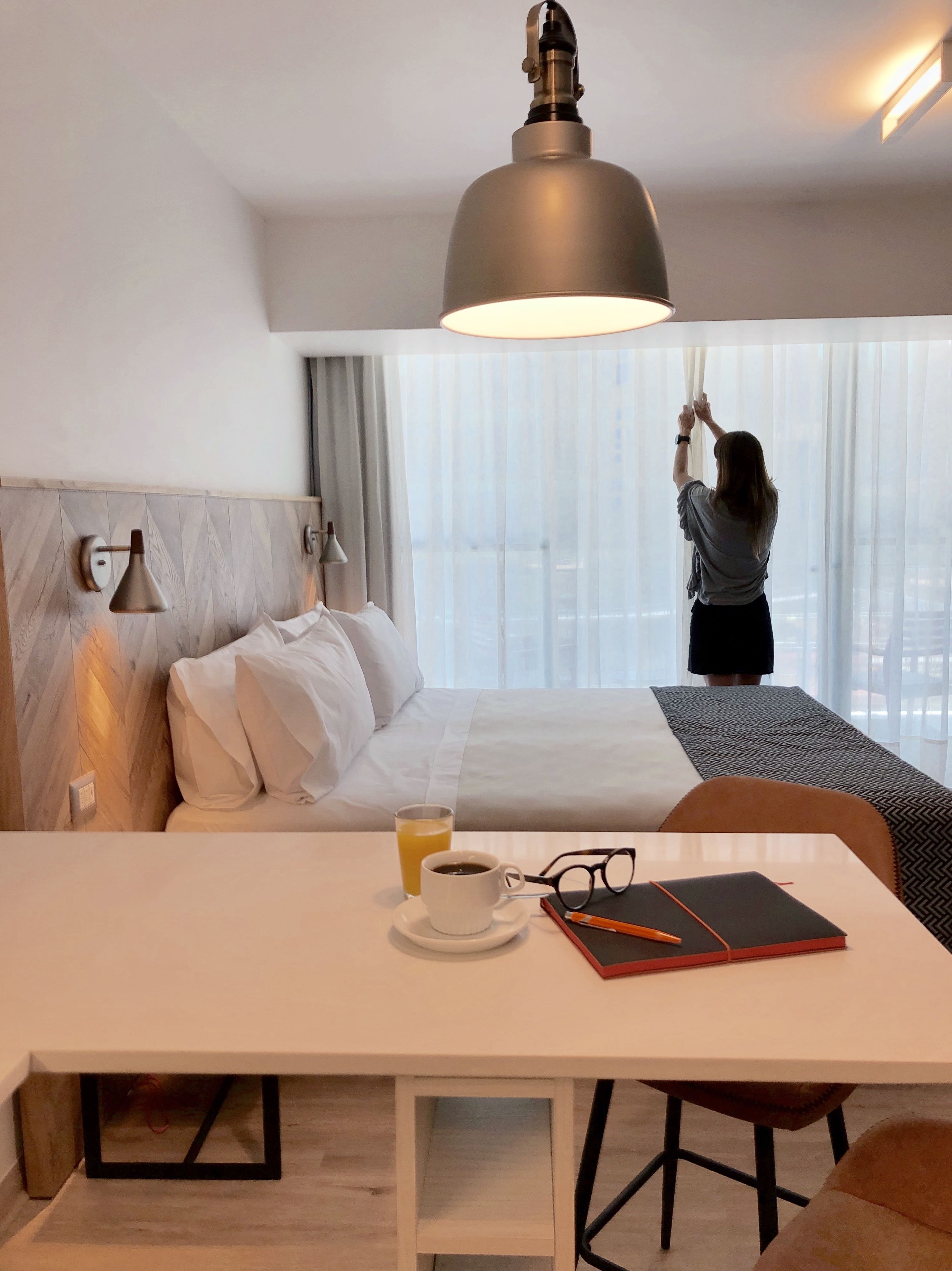 Una habitación de hotel con una cama, una mesa con una taza de café, un par de gafas y un cuaderno.