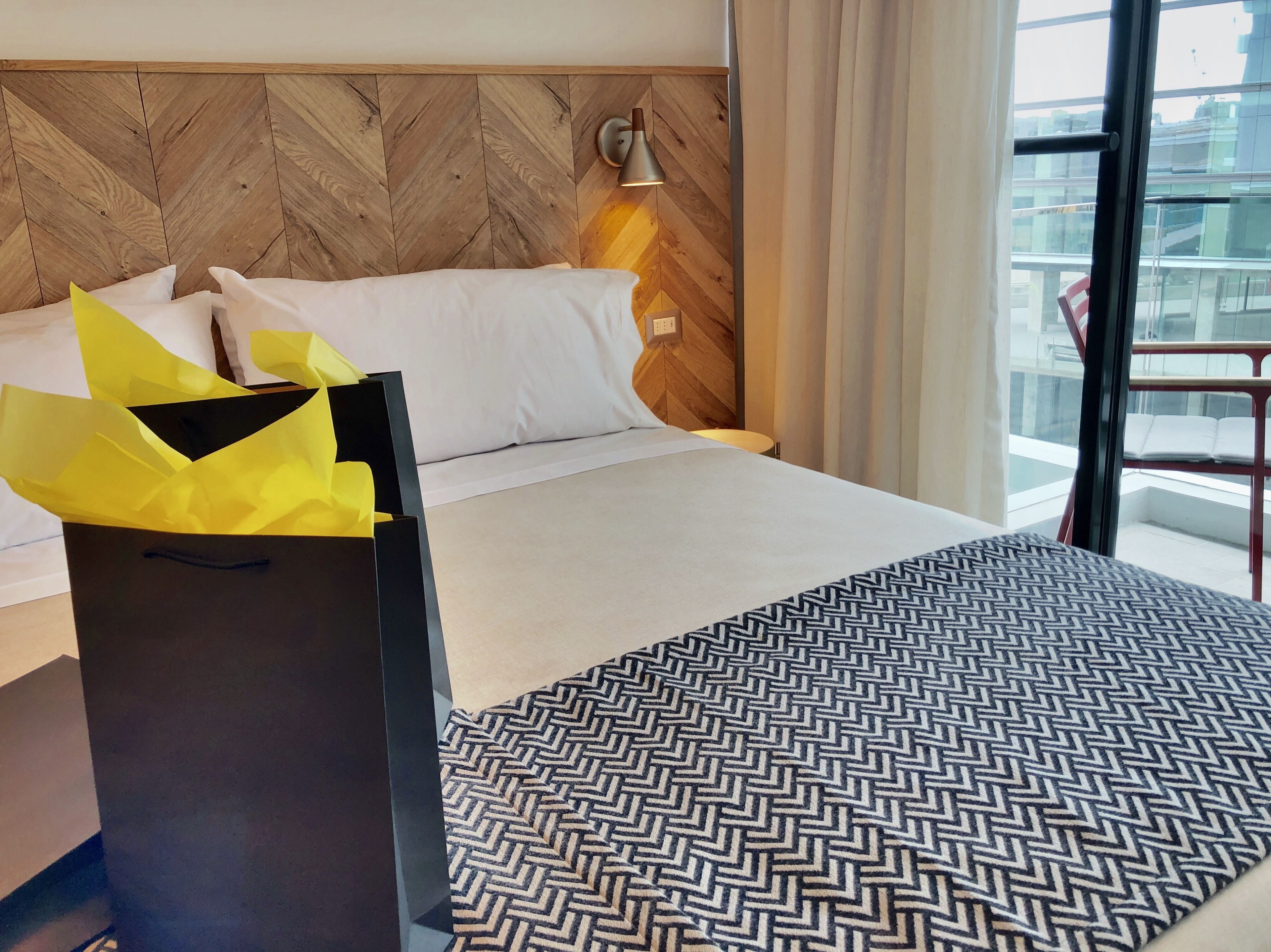 Habitación de hotel con cabecera de madera, cama con ropa blanca, manta con estampado y maleta negra con envoltorio amarillo.