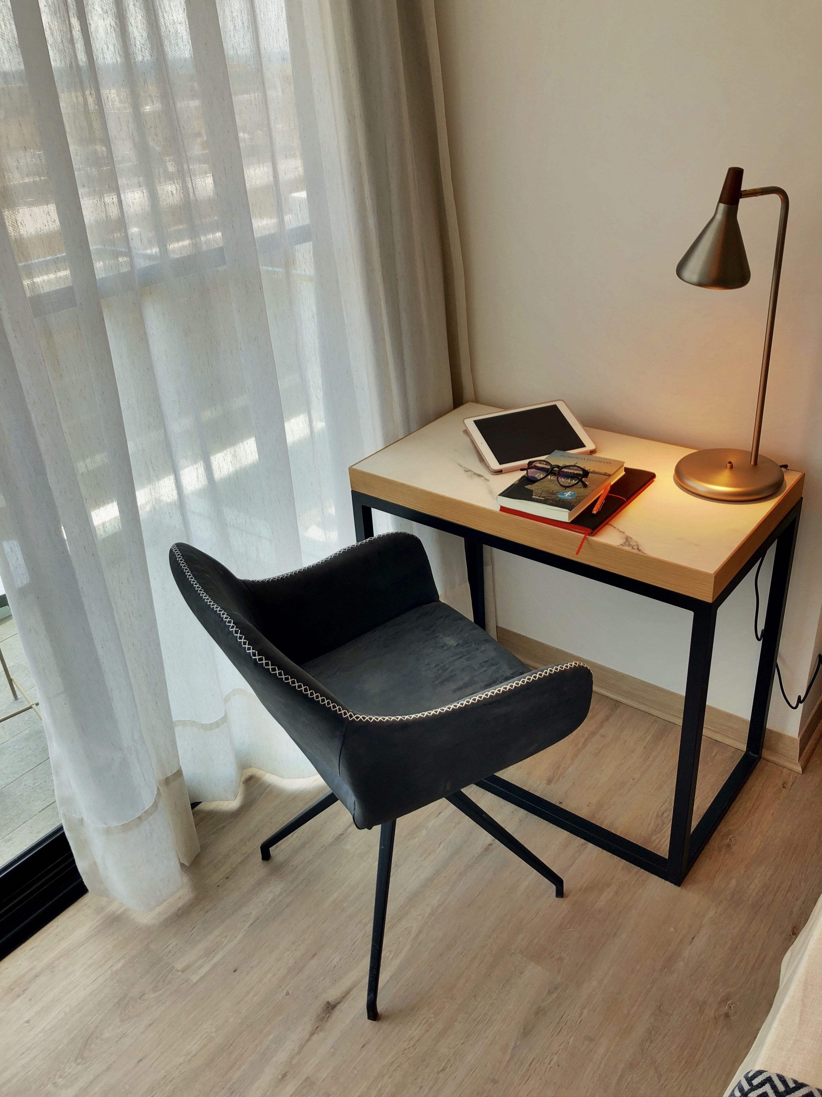 Quarto moderno com escrivaninha de madeira, cadeira preta, tablet, livro e luminária de chão.