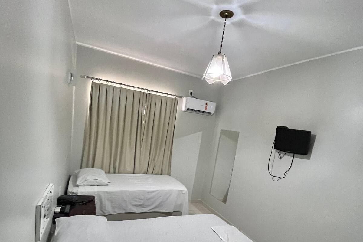 Quarto de hotel com duas camas, um ventilador de teto, um ar condicionado, uma televisão na parede e uma cortina.