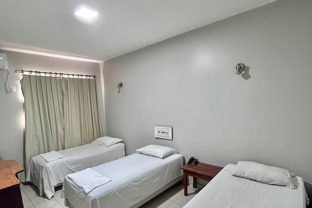 Quarto de hotel com duas camas de solteiro, uma escrivaninha, uma cadeira, mesa de cabeceira com abajur e janela com cortinas.