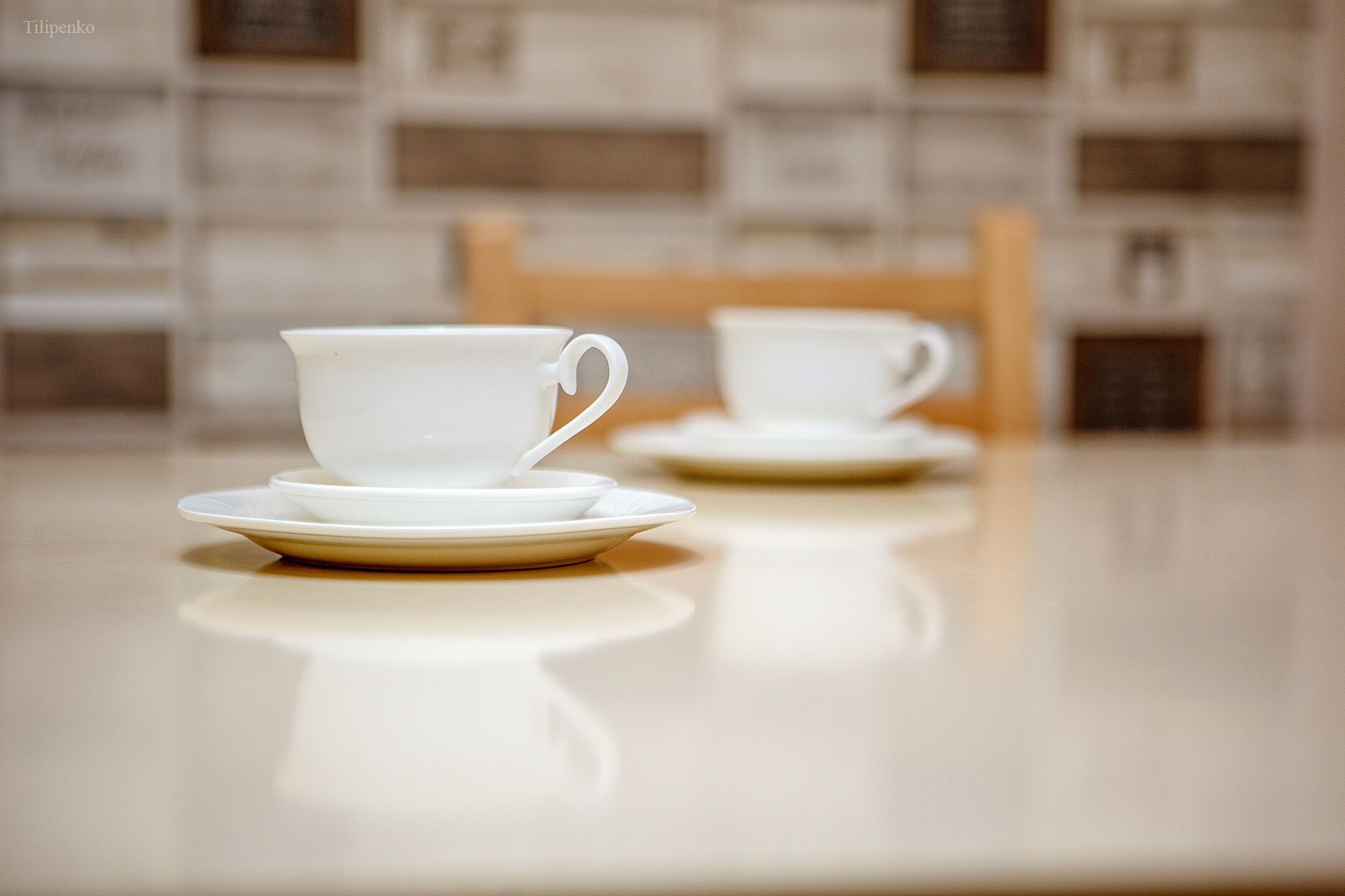 A table with two white cups and saucers, a wooden chair, and a tiled wall in the background.