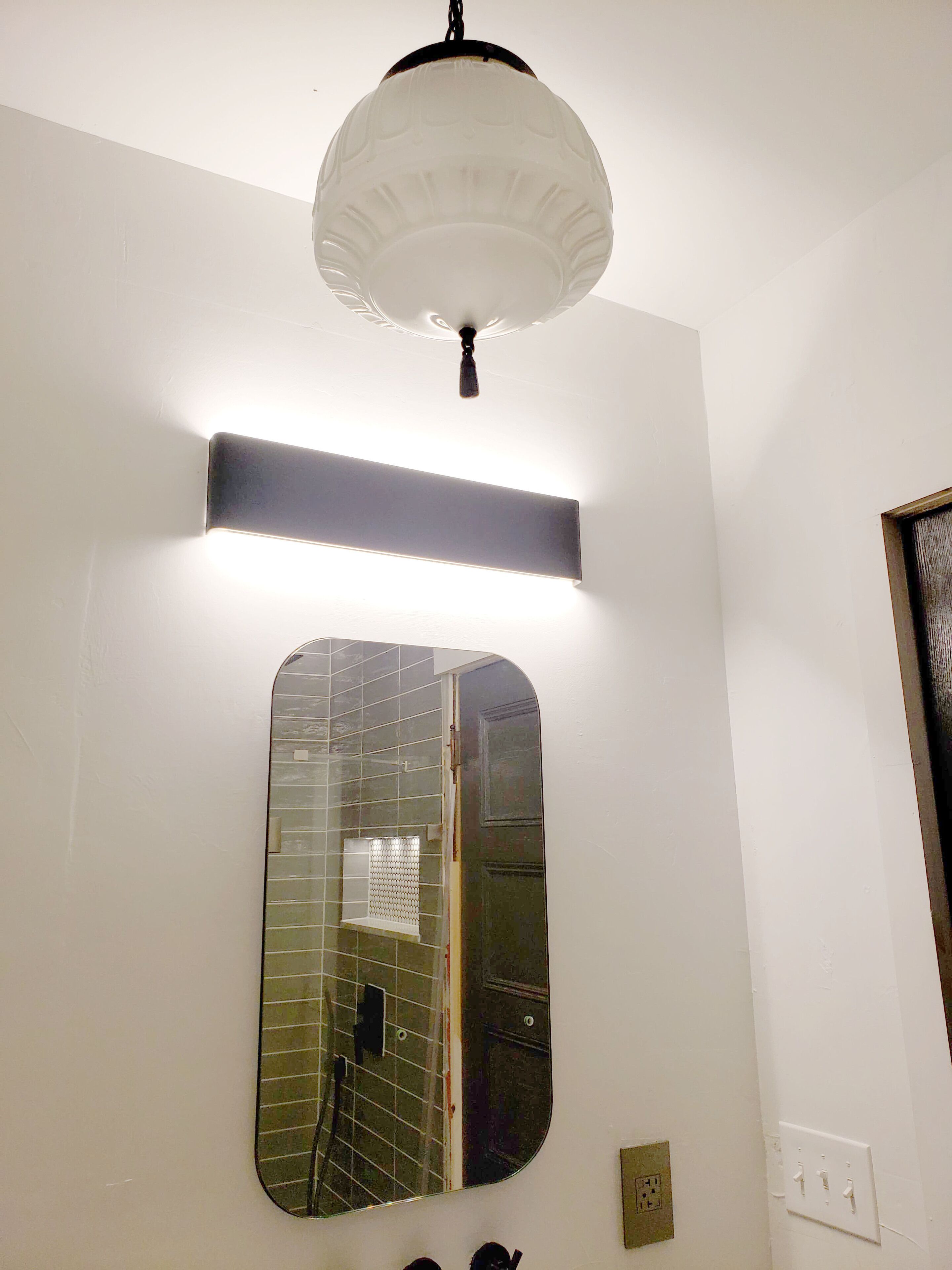 Une salle de bain avec un miroir, un luminaire et une porte.