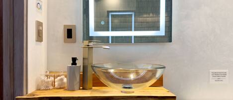 Un baño moderno con un lavabo de vidrio, un mueble de tocador de madera, un espejo y un letrero que indica no tirar nada más que papel higiénico.