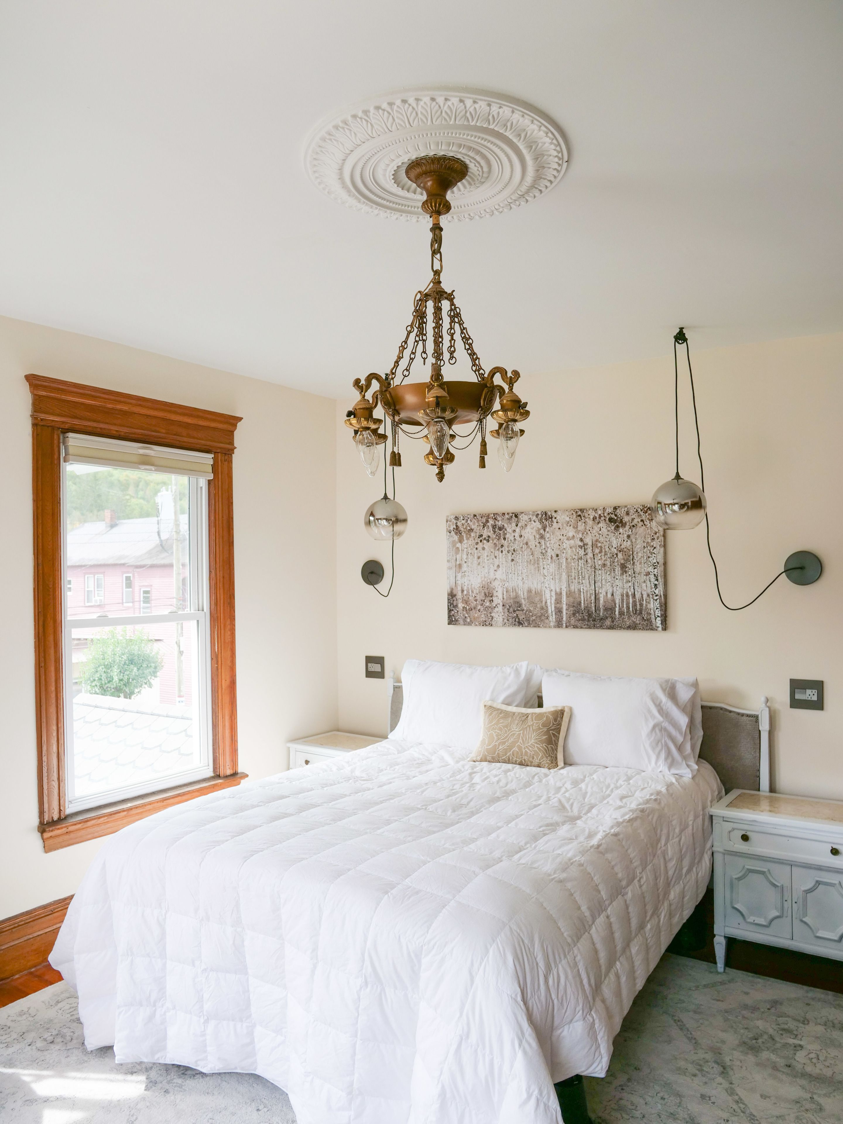 A bedroom with a large bed, a chandelier, a window, and a painting on the wall.
