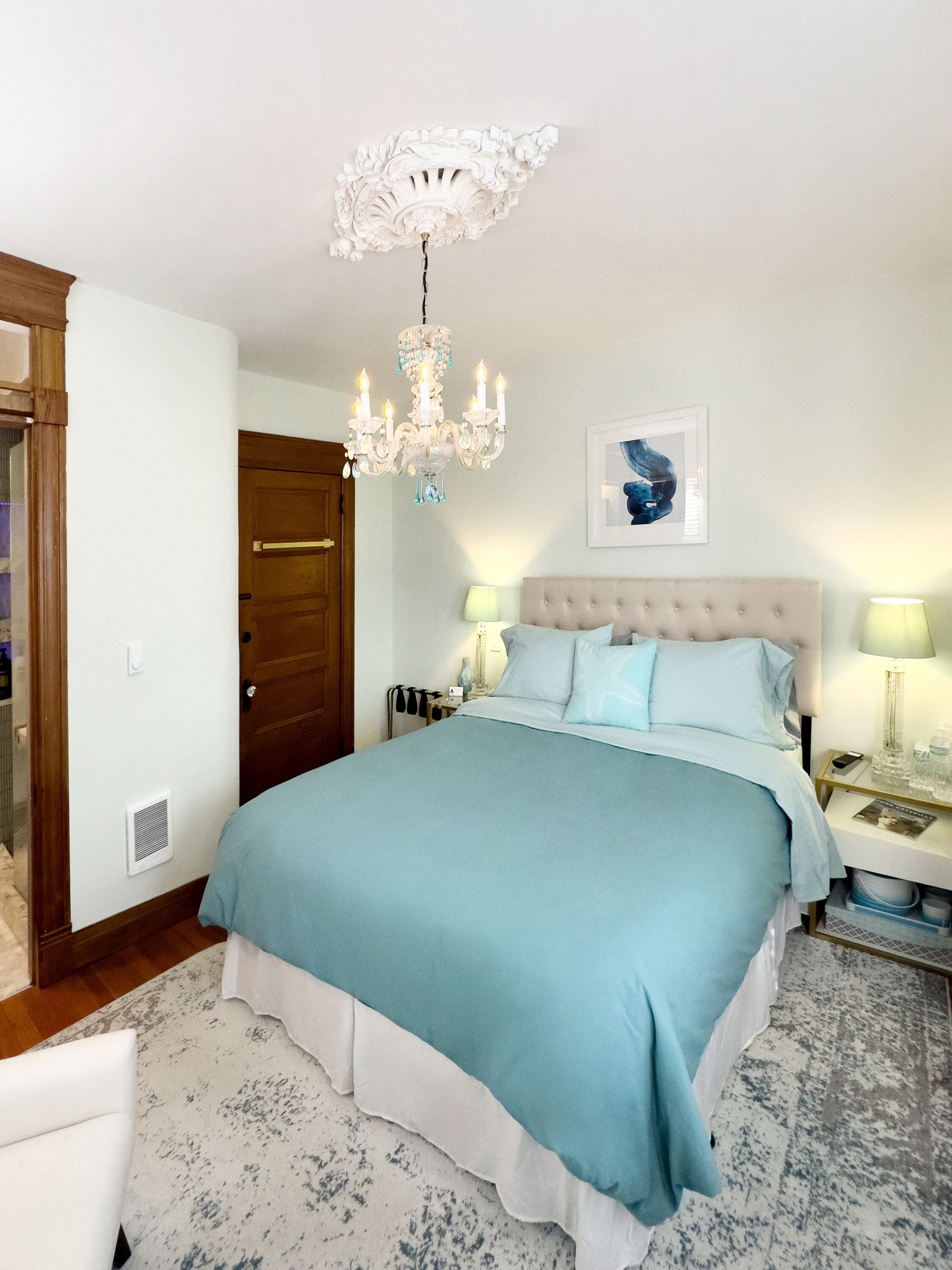 A bedroom with a large bed, a chandelier, a framed picture on the wall, and a wooden door.