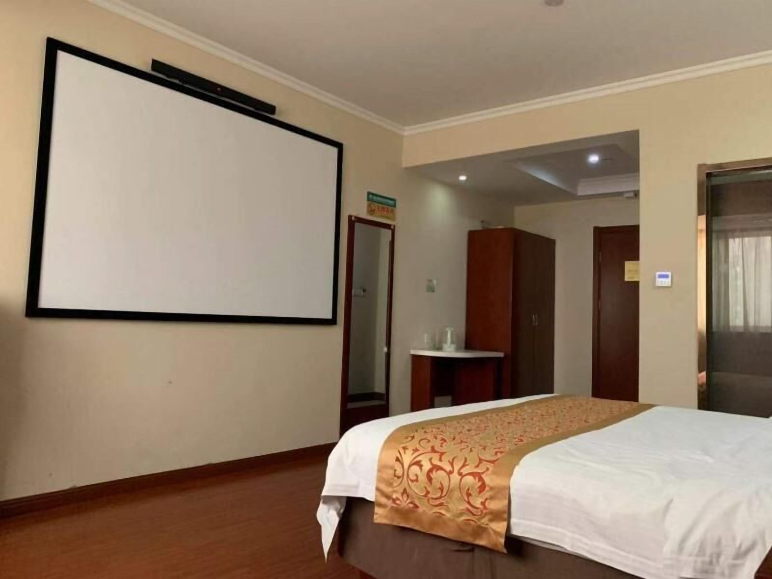 A hotel room with a large bed, a wooden floor, a white wall with a black-framed screen, a wooden wardrobe, and a small table with a glass top.