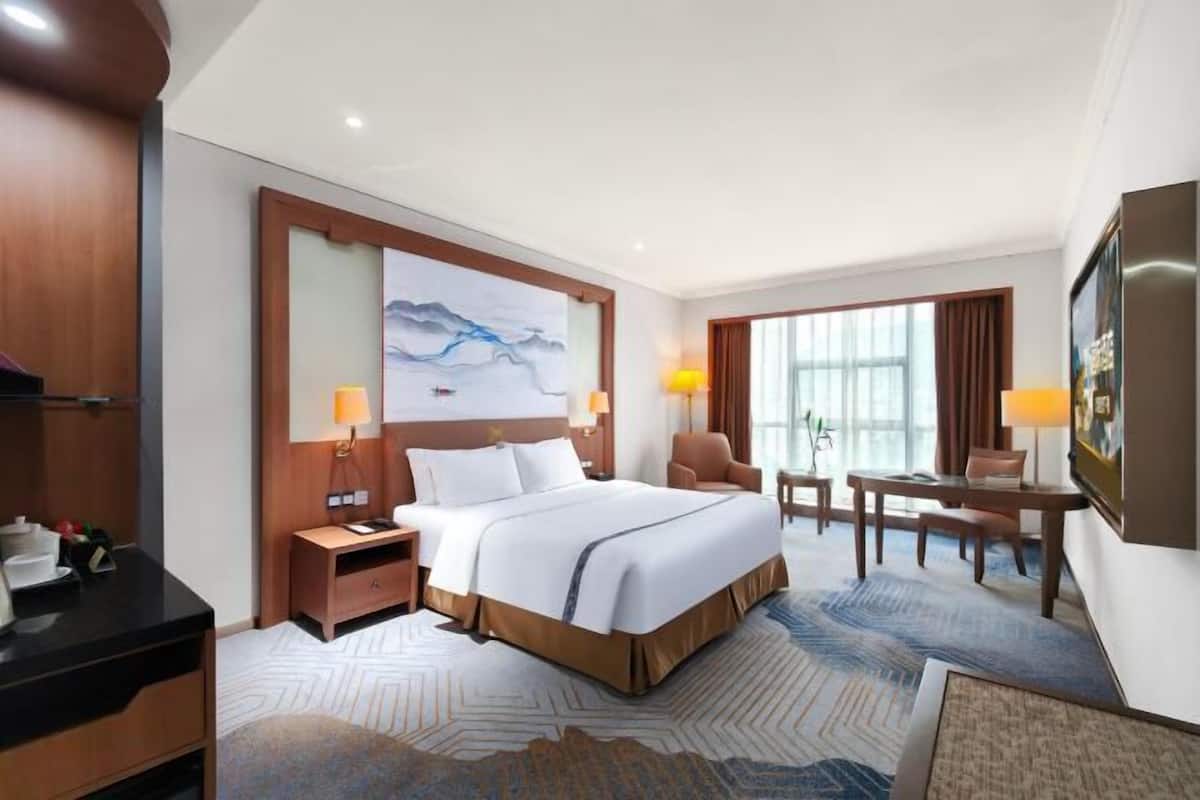 A hotel room with a bed, bedside tables, a desk, a chair, a TV, and a large window.