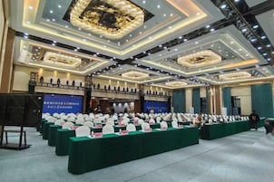 Un salón de conferencias con filas de mesas, un escenario y una pantalla grande.