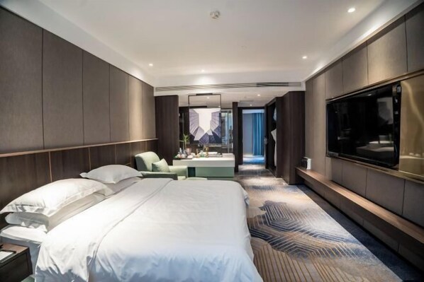 A modern hotel room with a large bed, a seating area, a TV, and a view of the city.