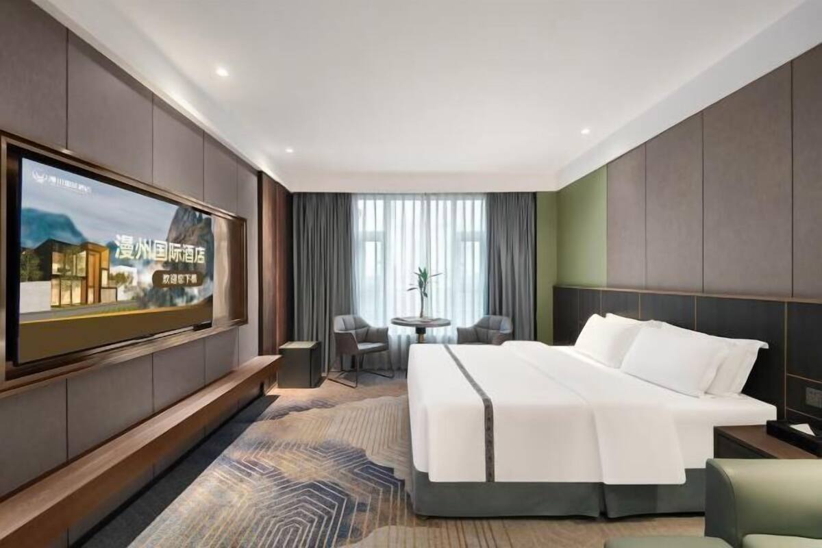 A hotel room with a large bed, a TV mounted on the wall, a small table, two chairs, and a window with curtains.