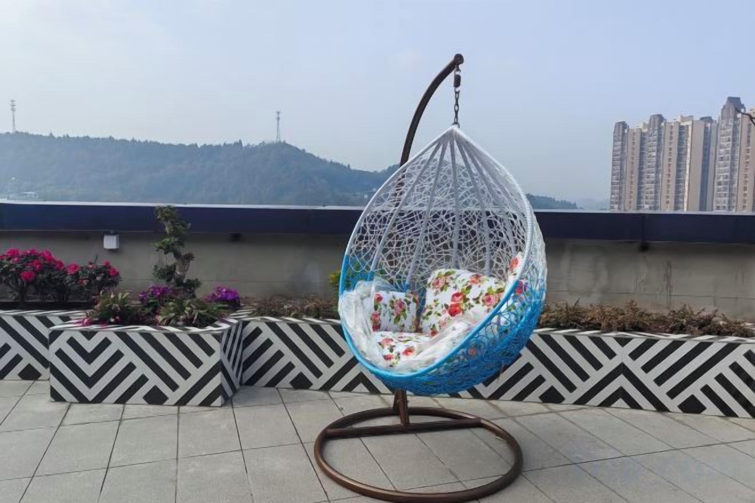 A hanging egg chair with a floral cushion on a rooftop terrace with patterned walls and a view of buildings and hills.