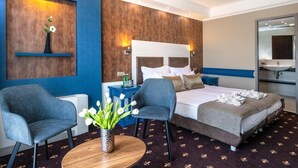 A hotel room with a bed, two armchairs, a small table with a vase, a nightstand, and a mirror. - Balneo Hotel Zsori Thermal Wellness (Mezőkövesd)