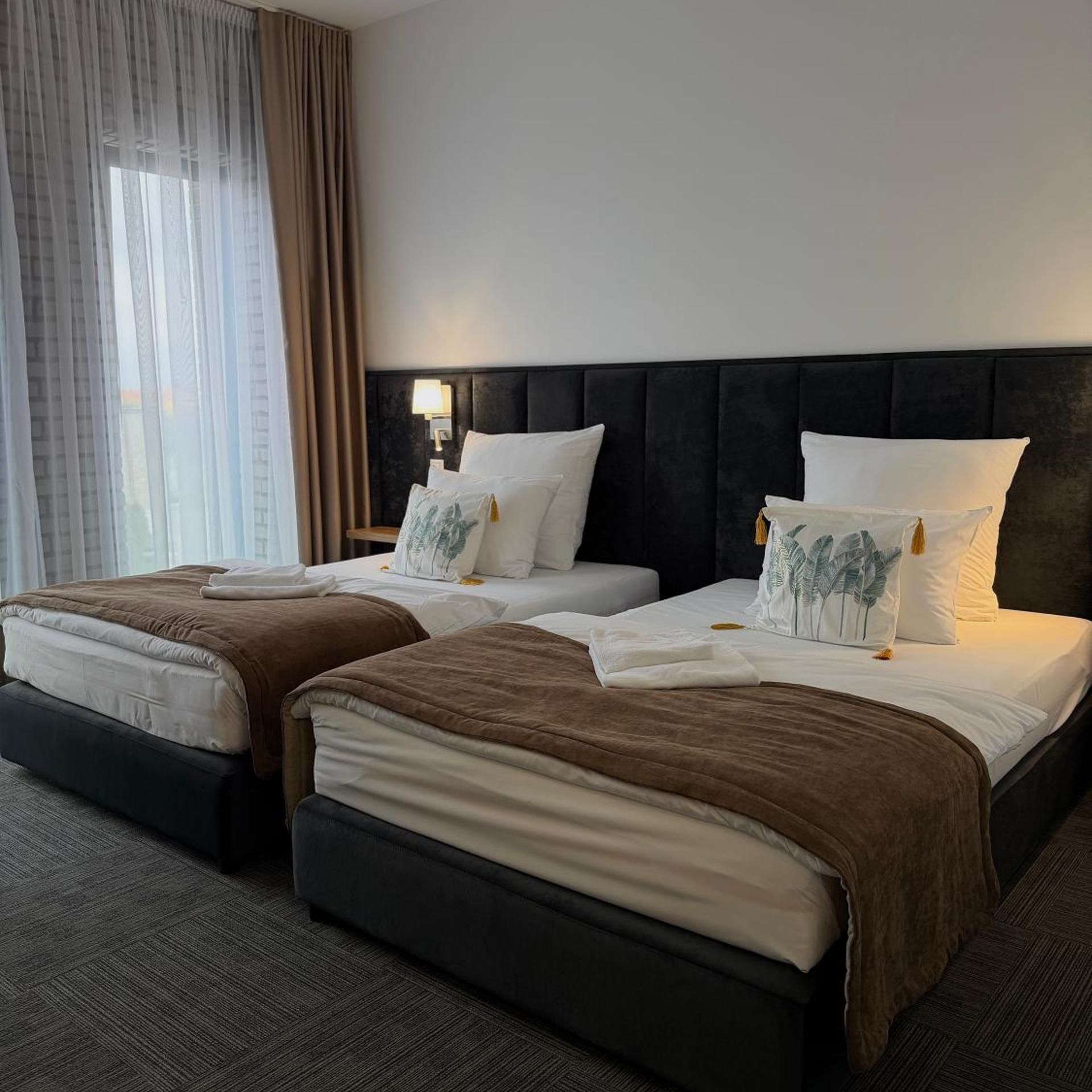 Ein Hotelzimmer mit zwei Betten, einem dunklen Kopfteil und einem Fenster mit durchsichtigen Vorhängen.