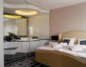 Een moderne hotelkamer met een gebogen glazen douchecabine, een ronde spiegel, een bed met hoofdbord en een nachtkastje met een lamp.