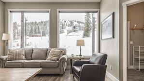 Un salon comprenant un canapé, un fauteuil et une table basse, de grandes fenêtres donnant sur un paysage enneigé, et un tableau encadré accroché au mur.