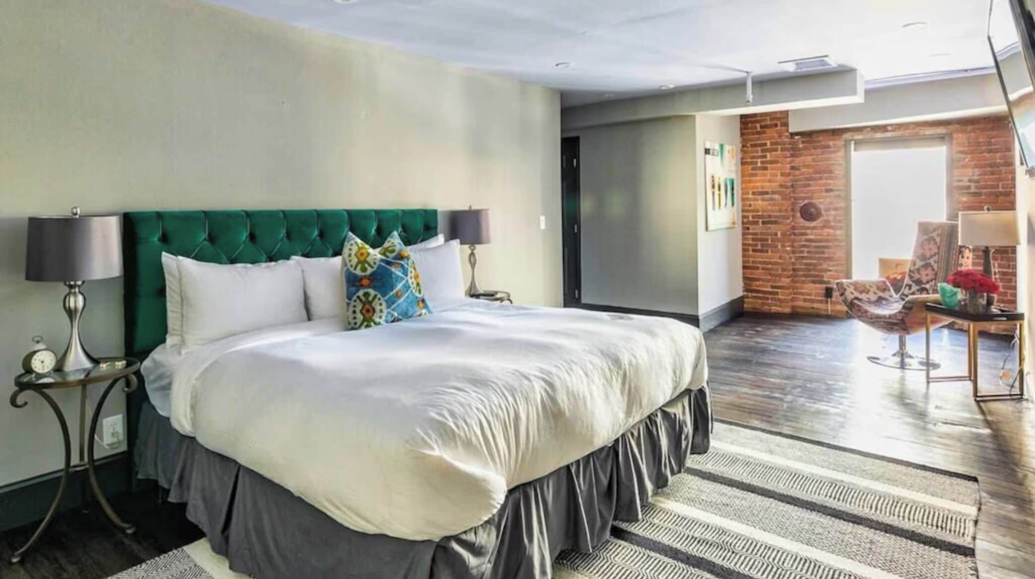 A modern hotel room with a large bed, a green upholstered headboard, and a view of a brick wall and a chair in the adjacent room.