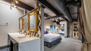 Une chambre d’hôtel moderne avec un meuble-lavabo en marbre, un grand lit recouvert d’une courtepointe foncée et un mur décoratif en briques.