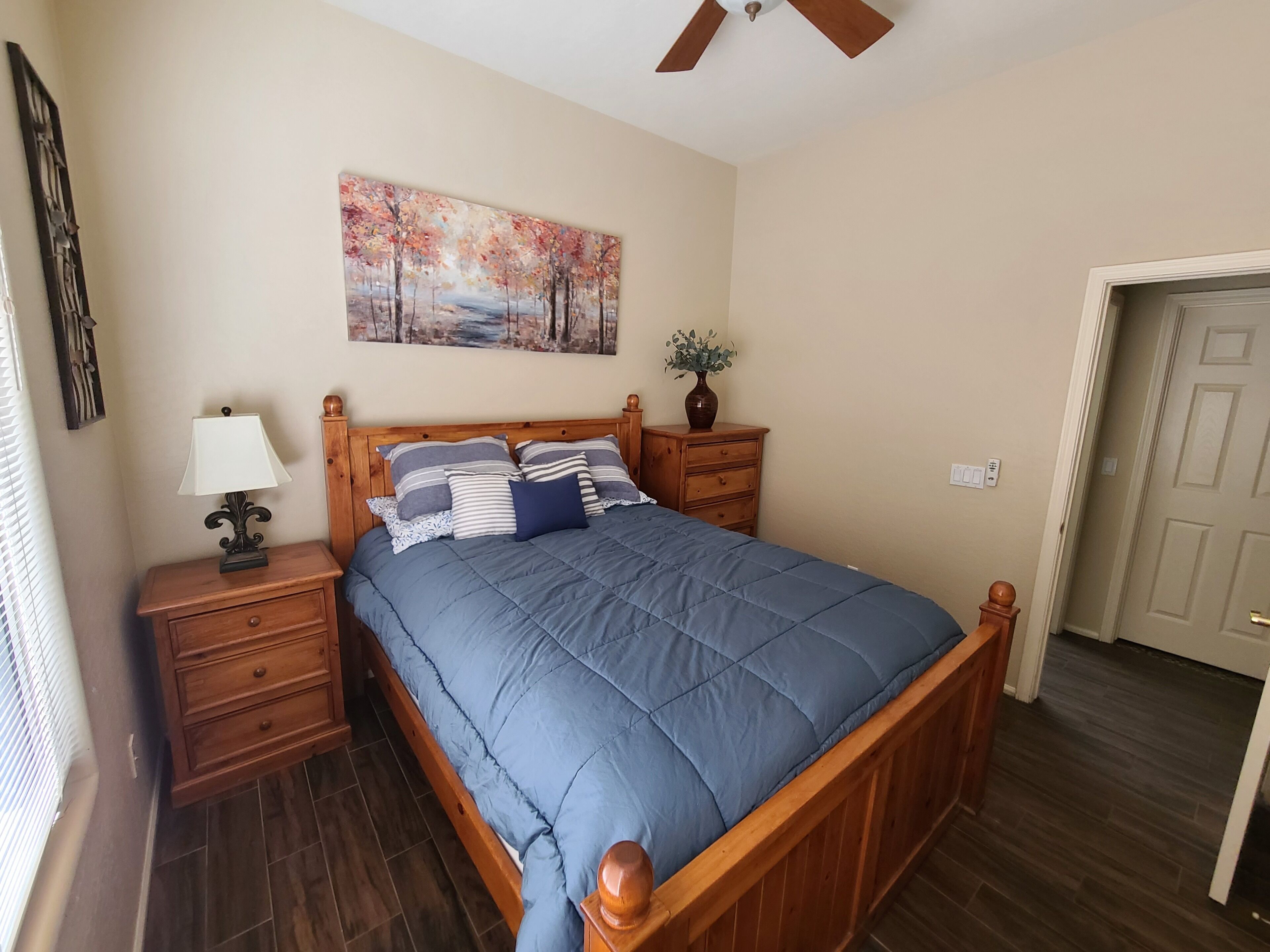 Quarto com cama de madeira, mesa de cabeceira com abajur, cômoda e um quadro na parede.
