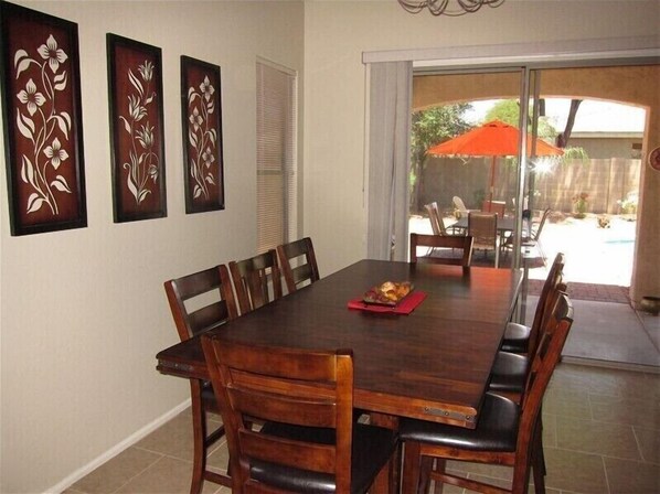 A dining area with a large wooden table and chairs, three wall-mounted floral decorations, and a sliding glass door leading to an outdoor patio with an orange umbrella.