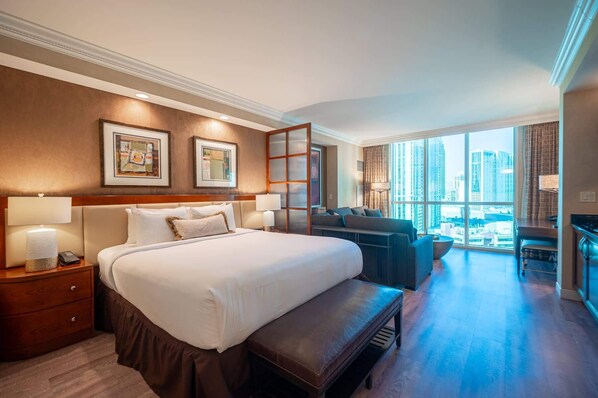 A hotel room with a large bed, a seating area with a sofa and armchairs, a desk, and a view of tall buildings through large windows. - No Resort Fee Strip View Suite + Free Valet+ Pool (Las Vegas)