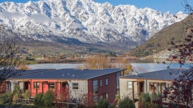 Une chaîne de montagnes dont les sommets sont coiffés de neige, un plan d’eau au premier plan et deux bâtiments à l’architecture moderne.