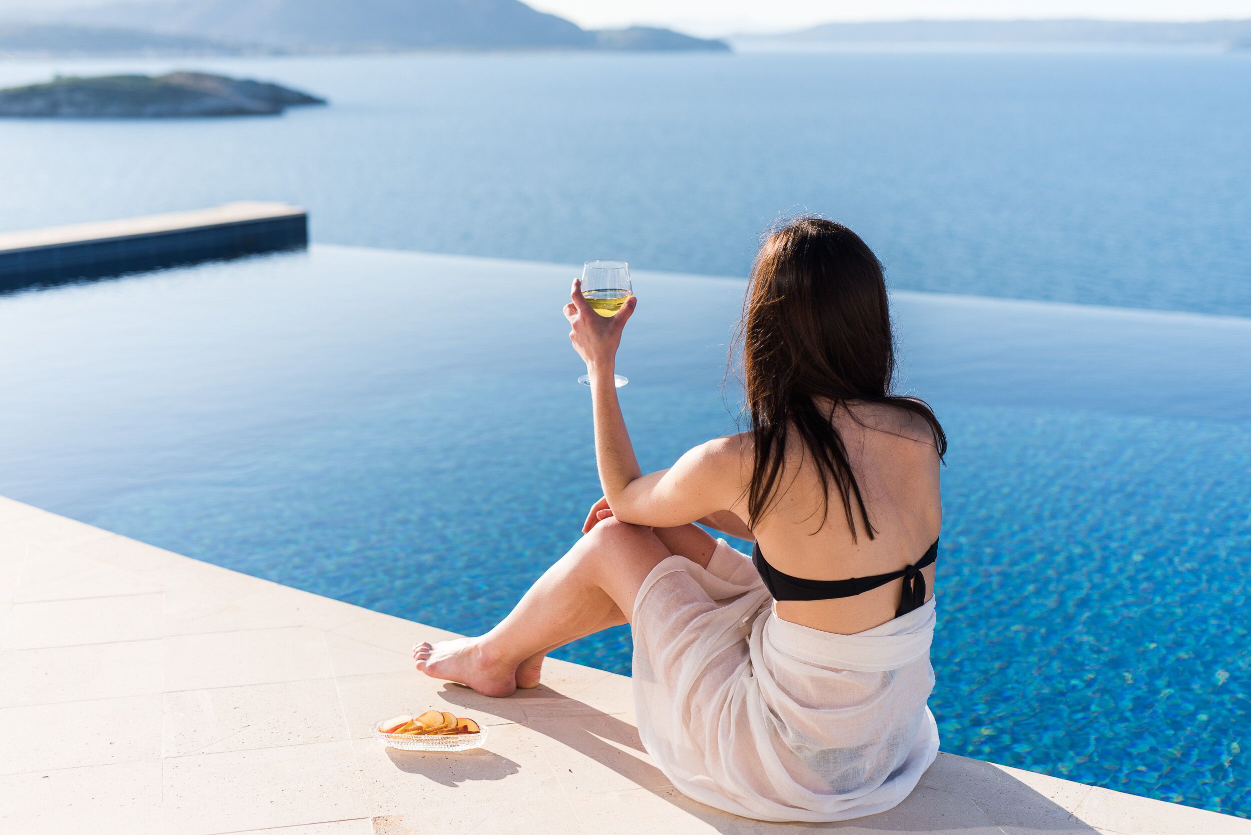 Une personne est assise au bord d’une piscine, un verre et une collation à portée de main, et elle a devant elle une étendue d’eau et une île lointaine.
