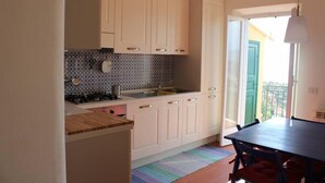 A kitchen with white cabinets, a gas stove, and a sink. There is a dining table with chairs and a colorful rug. - Apartment 6 pax wifi breathtaking sea view (Lerici)