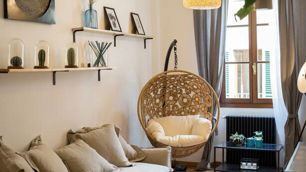 Una sala de estar acogedora con un sofá color beige, un sillón colgante, una mesita con plantas y una ventana con cortinas.