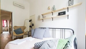 Un dormitorio con una cama, almohadas, una silla y estantes con objetos.
