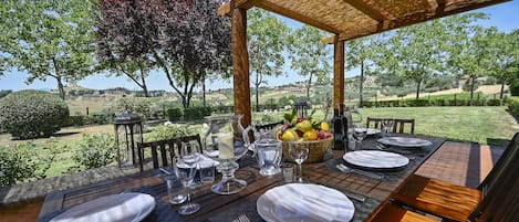 Un espacio exterior para comer con una mesa dispuesta para cuatro personas, rodeado de sillas de madera y una pérgola.