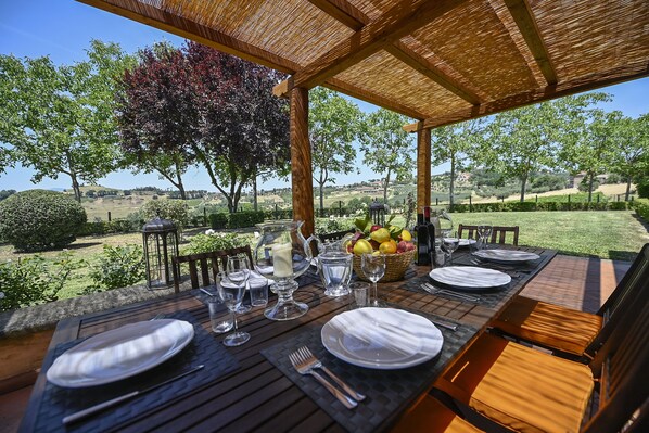Un espacio exterior para comer con una mesa dispuesta para cuatro personas, rodeado de sillas de madera y una pérgola.