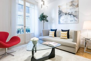 Ein modernes Wohnzimmer mit einem Glastisch, einem roten Sessel, einem grauen Sofa mit gemusterten Kissen, einer Topfpflanze und einem großen abstrakten Gemälde an der Wand.