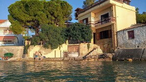 Un edificio costiero con una scala in pietra che conduce a una porta in legno, un balcone con ringhiera e un grande albero di fronte.
