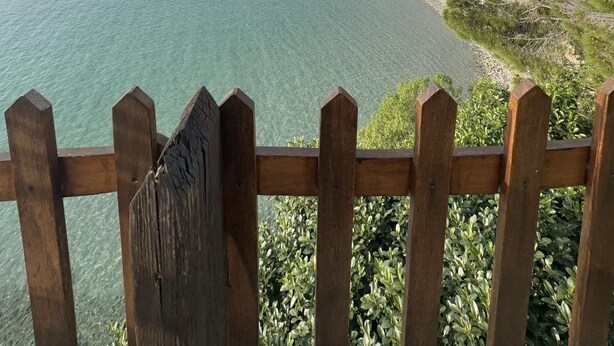Una vista sulla costa con una recinzione in legno, acqua turchese cristallina e una linea di costa lontana con edifici.