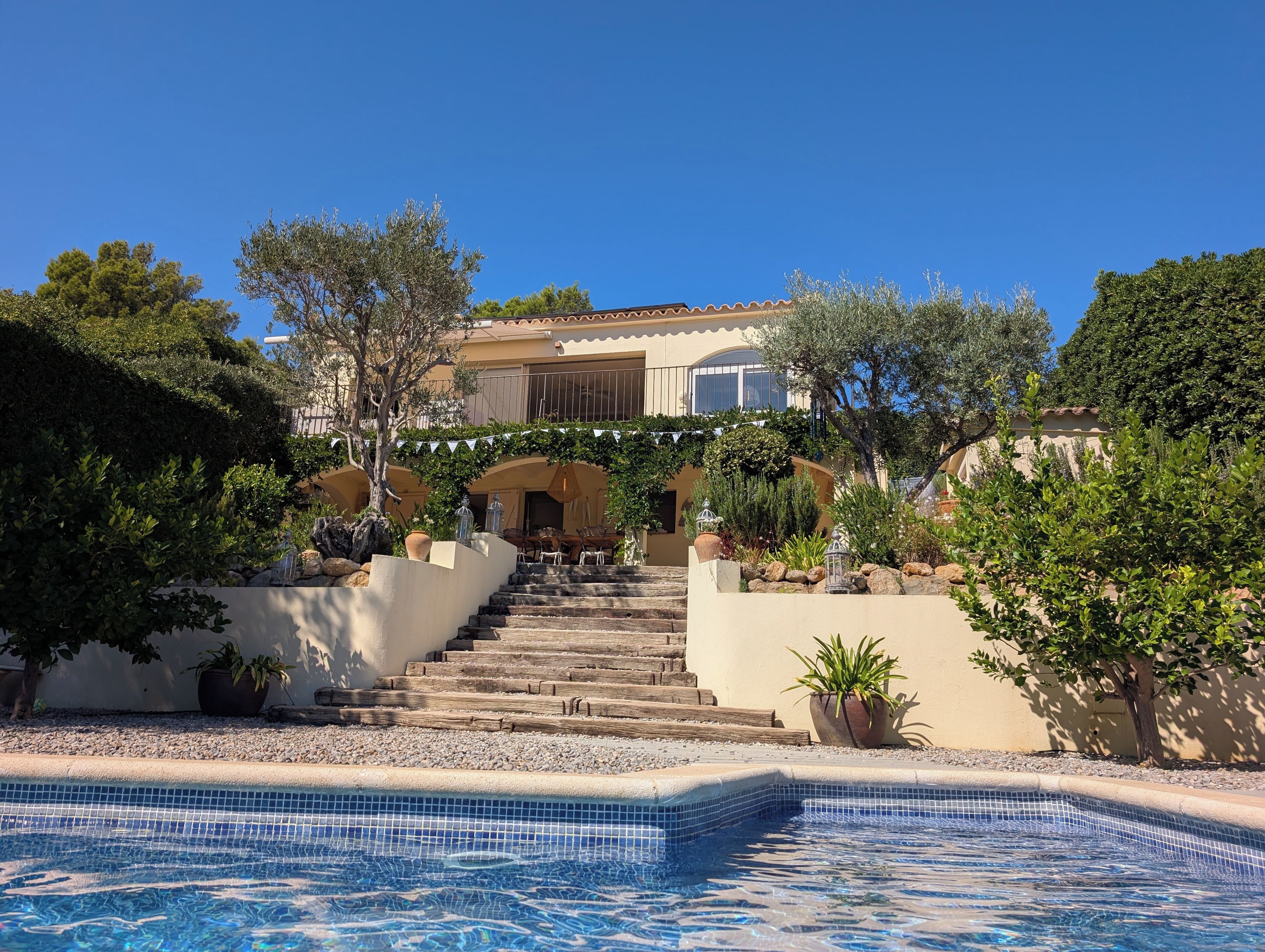 A pool area with a clear view of a building with a staircase leading to a door, surrounded by greenery and potted plants.