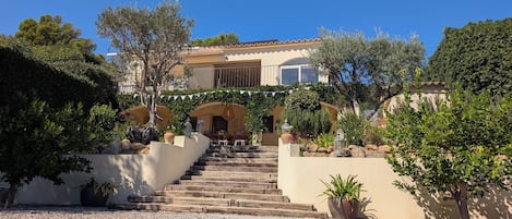 A pool area with a clear view of a building with a staircase leading to a door, surrounded by greenery and potted plants.