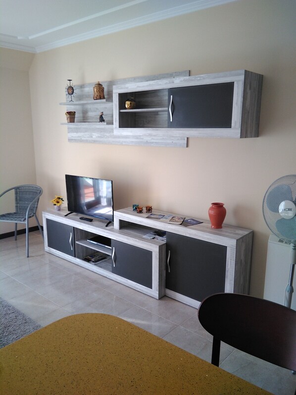 Una sala de estar con un televisor sobre un mueble bajo, un sofá, una silla y una mesita con adornos.