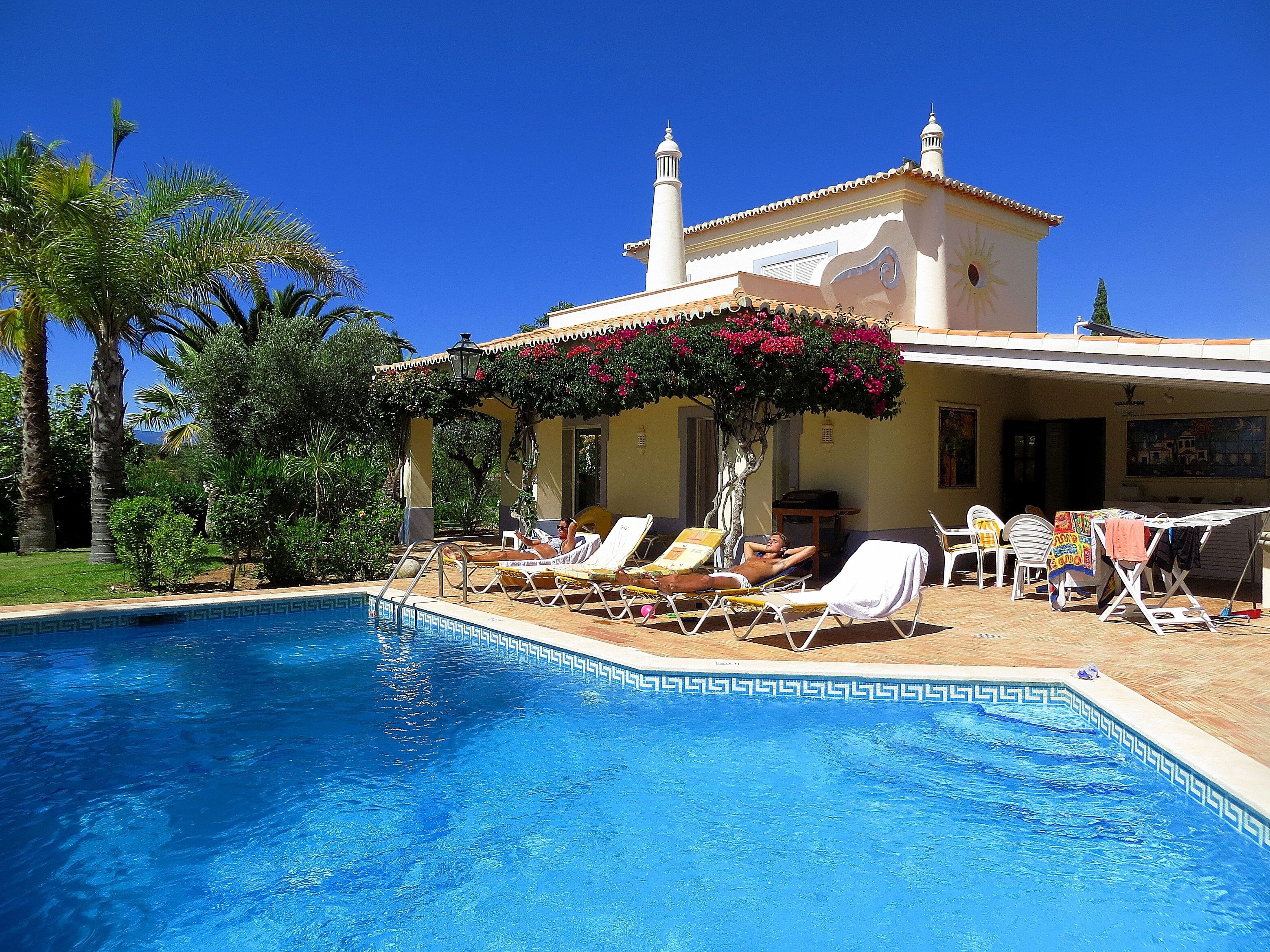 Un área junto a la piscina con sillones de descanso y una piscina, rodeada de palmeras y un edificio con techo de tejas.