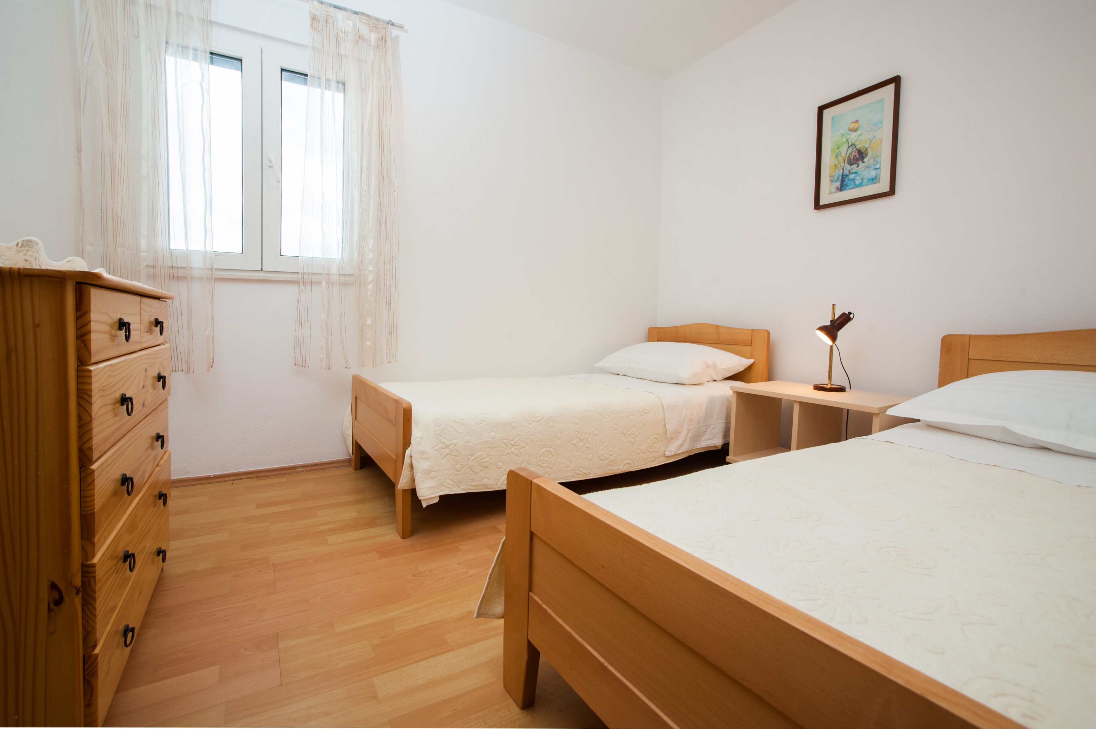 Una camera da letto con due letti singoli, una cassettiera in legno, un comodino con una lampada e una finestra con tende leggere.