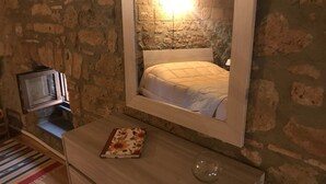 A bedroom with a stone wall, a bed with a white headboard, a bedside table with a book and a glass, and a TV on a stand.